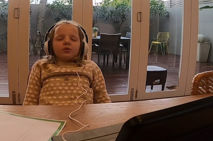 A six-year-old girl wears headphones and has her eyes closed sitting in front of a computer.