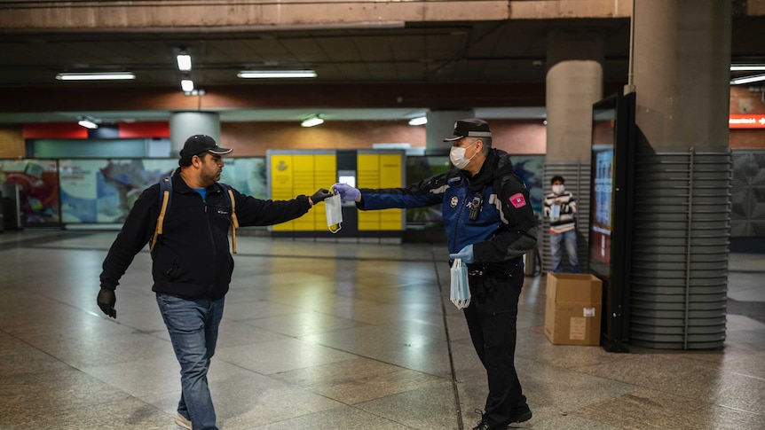 Police officer hands man a mask inside a train station. Both men have their arms outstretched to keep distance