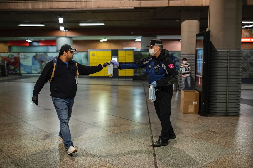Police officer hands man a mask inside a train station. Both men have their arms outstretched to keep distance