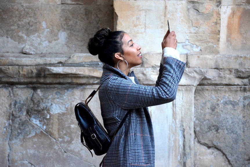 A woman wearing a jacket takes a photo on her phone in front of a granite or sandstone wall.