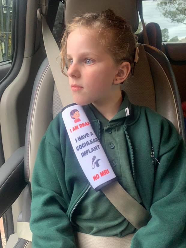 A girl sits in a car seat looking out the window with a seat belt cover with medical information attached to her seat belt.