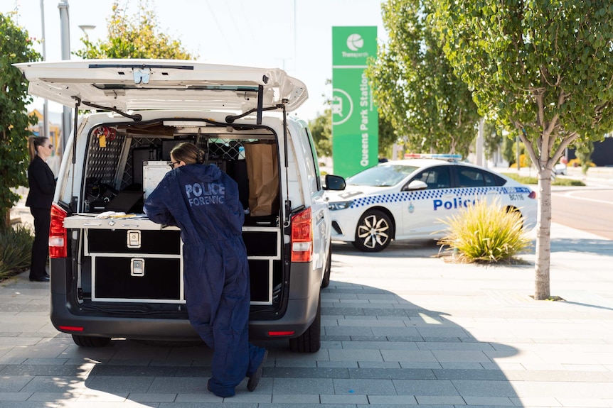 A WA Police forensic officer stands at the back of a police van near another police car outside Queens Park train station.