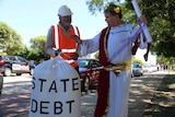 A man dressed as an emperor in robes and another in a hard hat holding a bag marked "state debt" stand on a street.