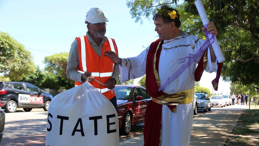 A man dressed as an emperor in robes and another in a hard hat holding a bag marked "state debt" stand on a street.