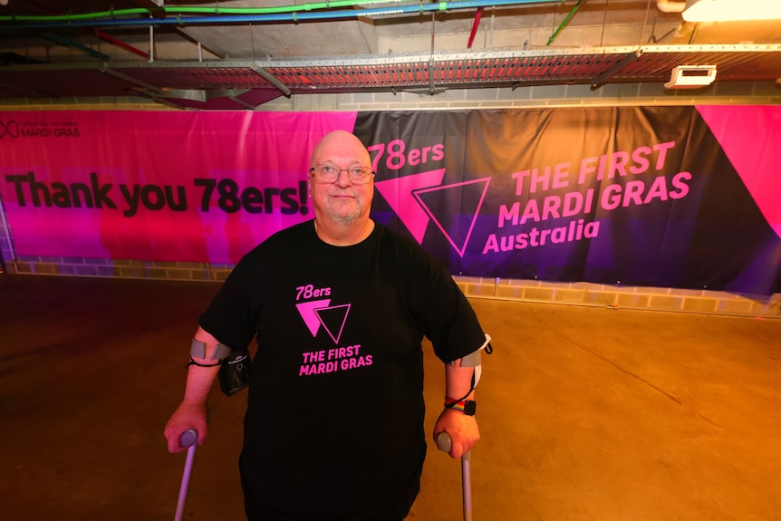 Peter stands with a mobility aid secured to each arm, wearing a 78ers shirt and in front of a banner thanking 78ers.