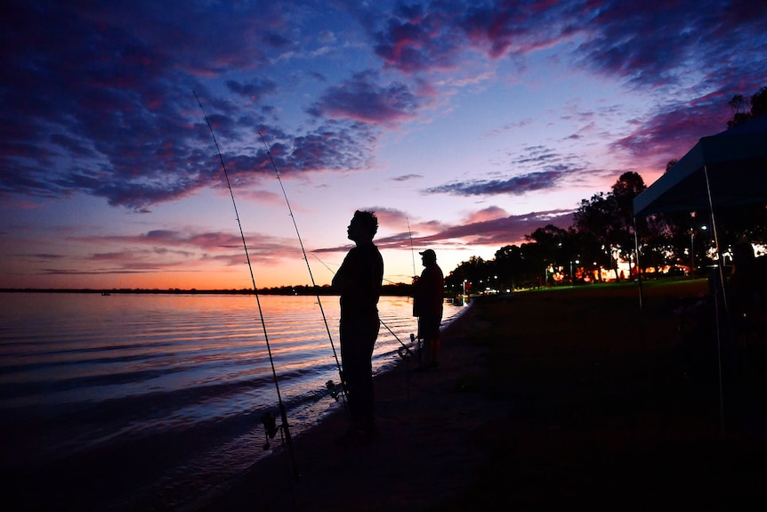 A man with fishing rots at a lake at sunrise.