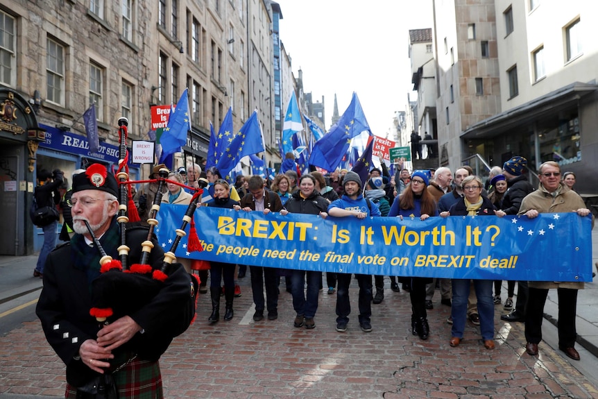 A piper leads a group of protesters who are holding a banner with the word Brexit is it worth it on it