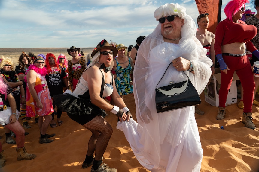 A man drressed as a bride, another in drag behind him, holding the bridal train with a crowd on the sand dune looking on 