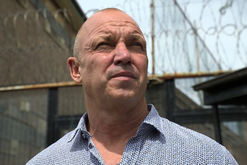 Close shot of a bald man in blue collared shirt with barbed wire behind