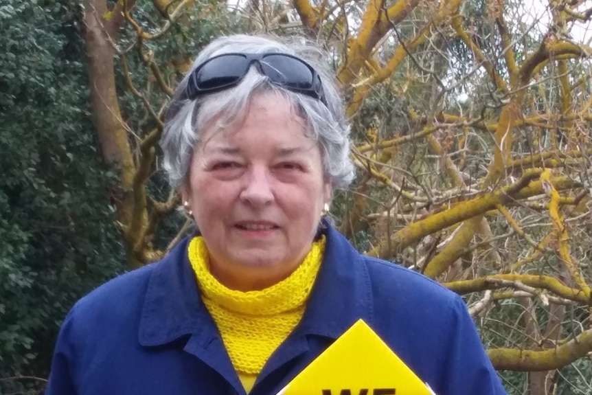 A woman with short, grey hair and sunglasses smiles at the camera while holding a yellow protest sign.
