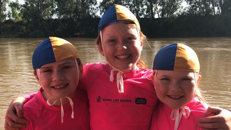Three girls in pink swim shirts and yellow and blue swim caps.