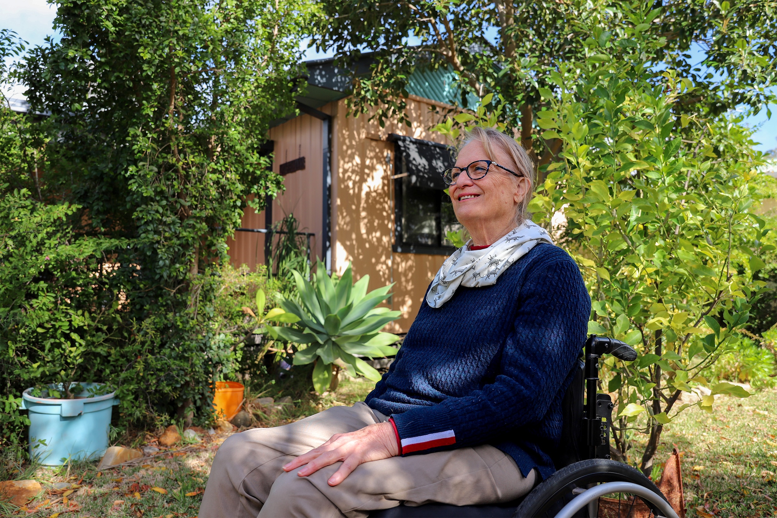 A woman sits in a wheelchair in a garden. She is smiling and wearing a blue jumper and a white scarf around her neck.