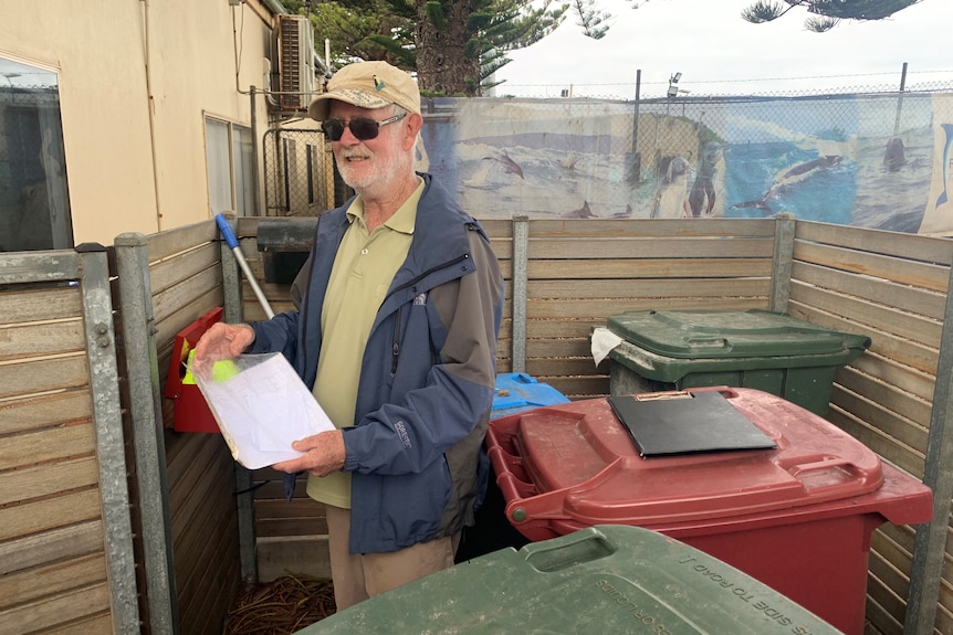 A man stands in a fenced area with several bins and a repurposed letterbox