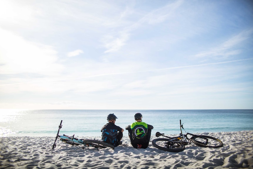 Two male mountain bike riders sitting on a beach with their bikes, looking out to the ocean.