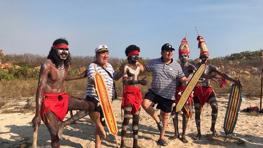 Two white male tourists pose with Aboriginal dancers on a beach.