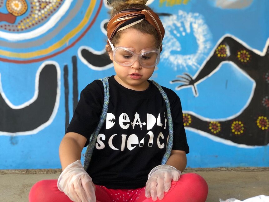 A young girl wears a Deadly Science t-shirt, disposable gloves and protective goggles in front of a colourful wall