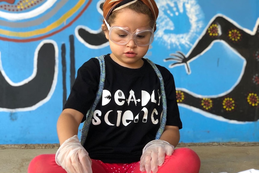 A young girl wears a Deadly Science t-shirt, disposable gloves and protective goggles in front of a colourful wall
