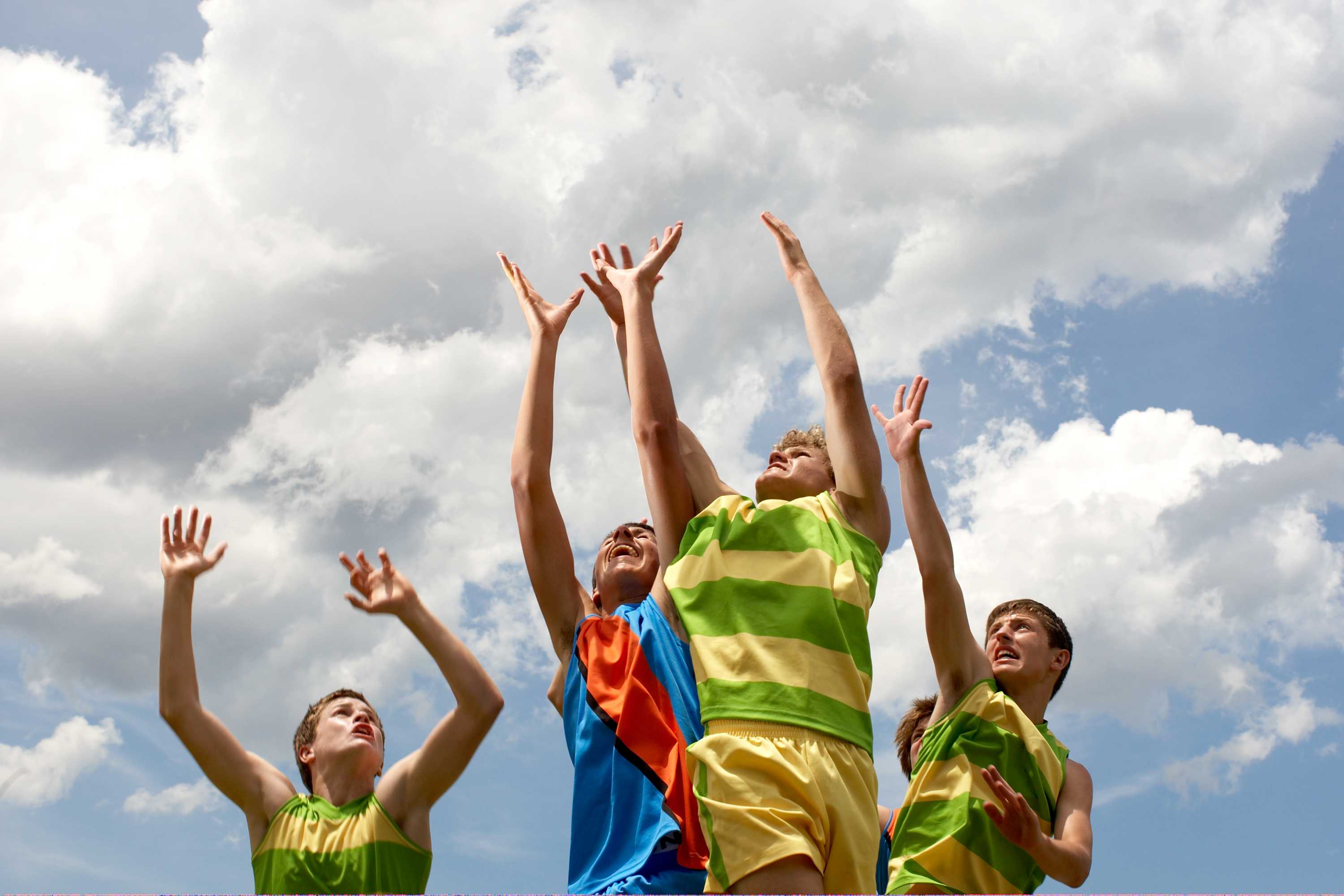 Four teenage boys leaping into the air as if to catch a ball, wearing sports jerseys.