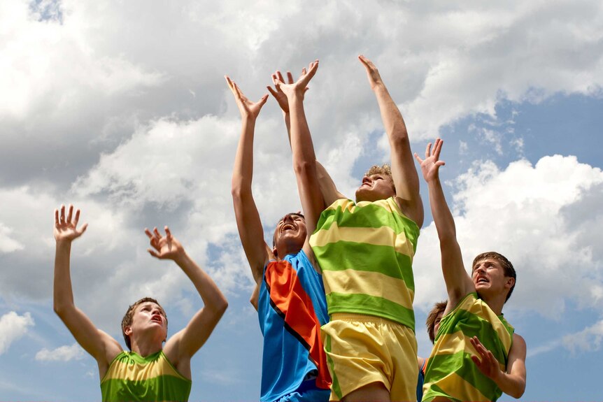 Four teenage boys leaping into the air as if to catch a ball, wearing sports jerseys.