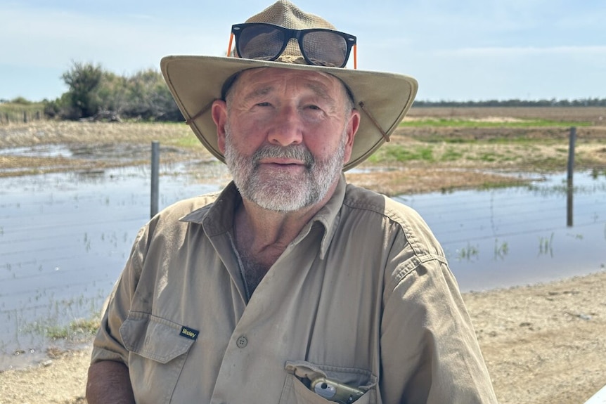 A man, wearing a farm shirt and a hat, stands in front of a flooded paddock.