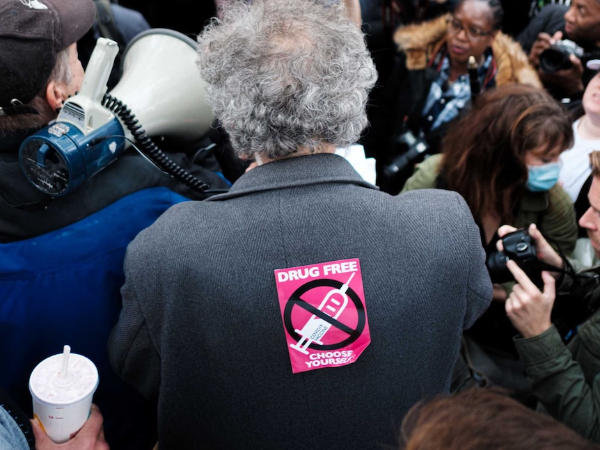 Man in grey jacket and a bright pink sticker on his back that says 'drug free choose yourself' in front of a crowd