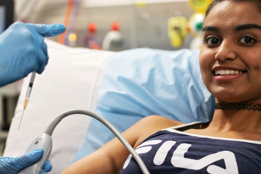 Close-up of a teenage girl lying against a pillow while gloved hands hover nearby with an injection.