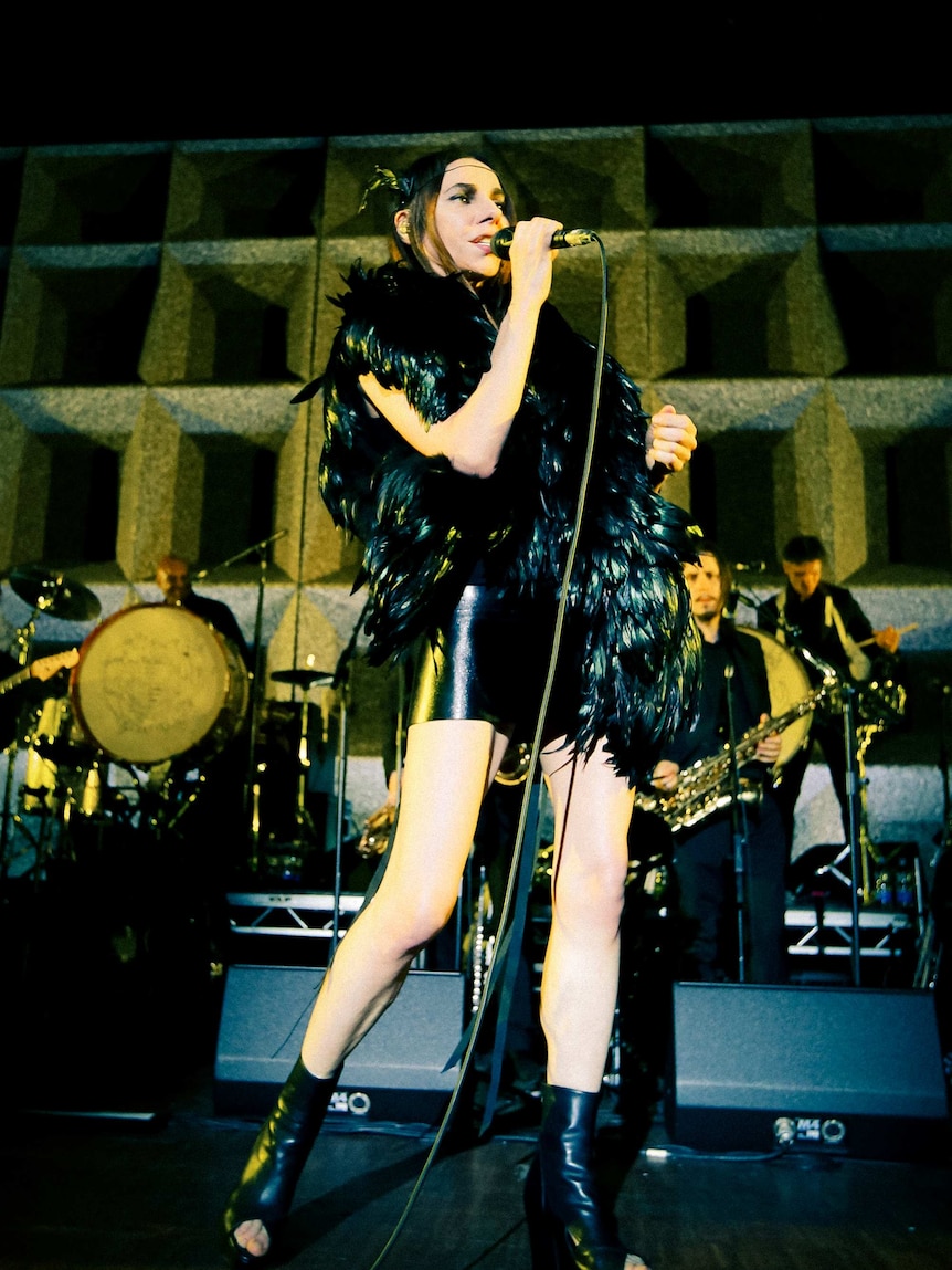 PJ Harvey performs with her band wearing black leather boots and feathers.