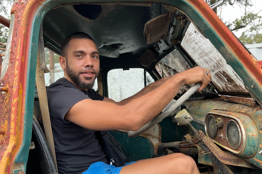 A young Indigenous man sits behind the wheel of an old rusty fire truck.