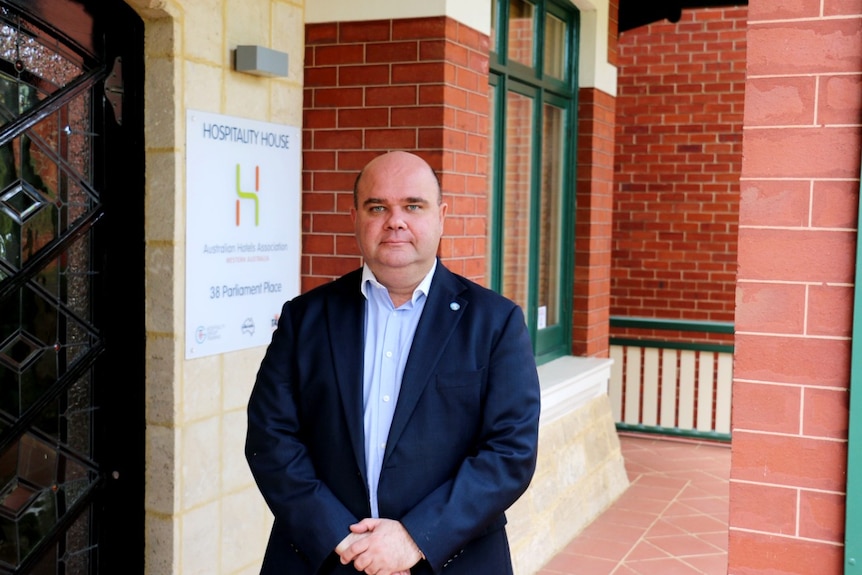 A man wearing a navy blazer and open shirt stands with his hands clasped in front of a brick wall.