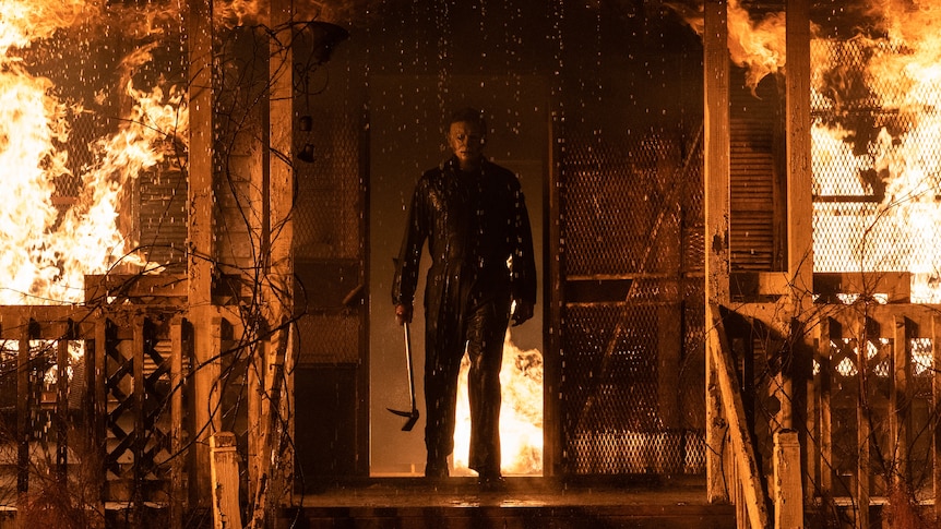 A man in a mask in overalls covered in blood and holding an axe stands in the doorway of a burning house