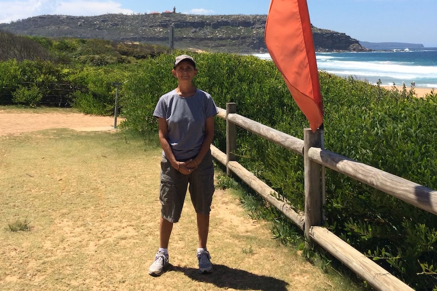 A woman in a shirt, short and caps stands next to a wood barrier by the beach.