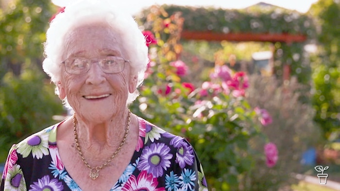 Elderly lady smiling at camera, standing in a garden filled with flowers