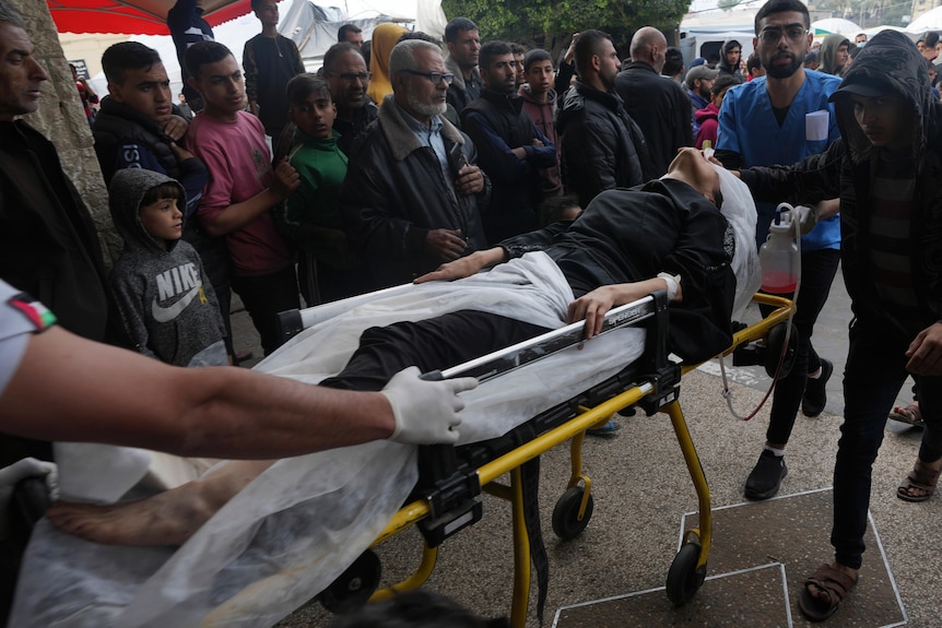 A bandaged woman on a hospital stretcher is pulled through a crowd of people outdoors.