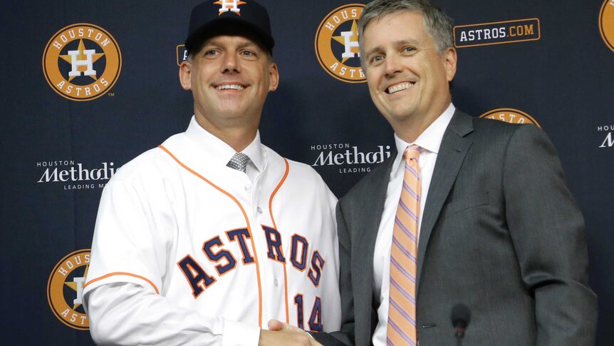 An older man wears an Astros jersey over a shirt and tie and an Astros cap, shaking hands with a man in a suit.