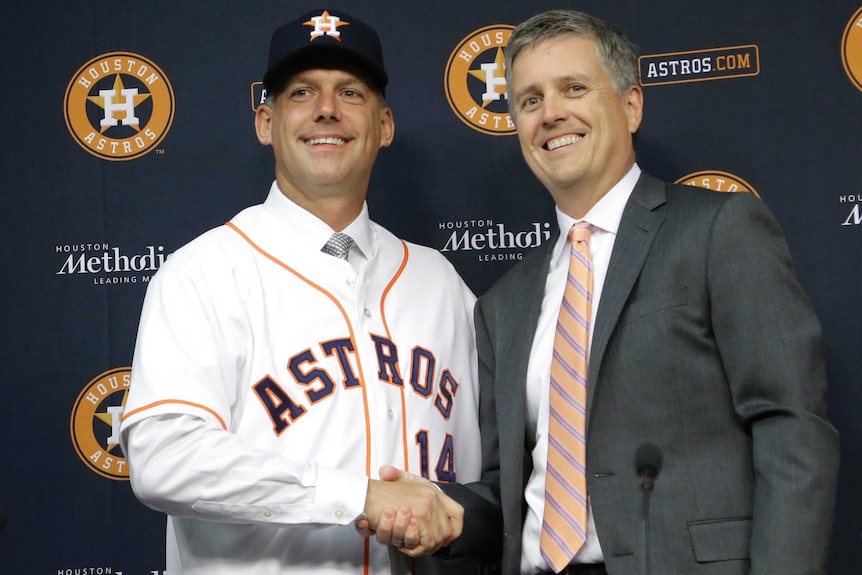 An older man wears an Astros jersey over a shirt and tie and an Astros cap, shaking hands with a man in a suit.