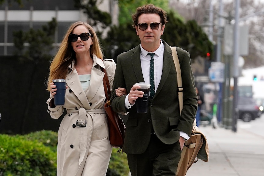 A man and woman in corporate wear with large sunglasses walking down a street holding coffee. 