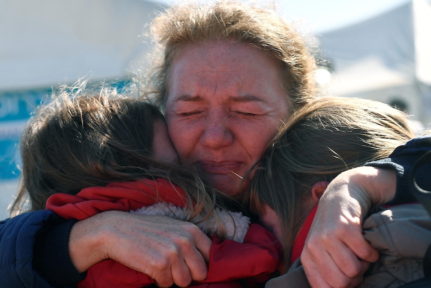 A woman hugs two people whose heads are facing away from the camera as she cries.