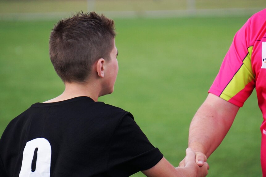 A young boy in a play playing guernsey shakes hands with a larger man in a pink playing shirt.