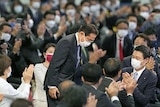 Fumio Kishida wearing a mask bows while standing on a platform surrounded by a crowd of people. 