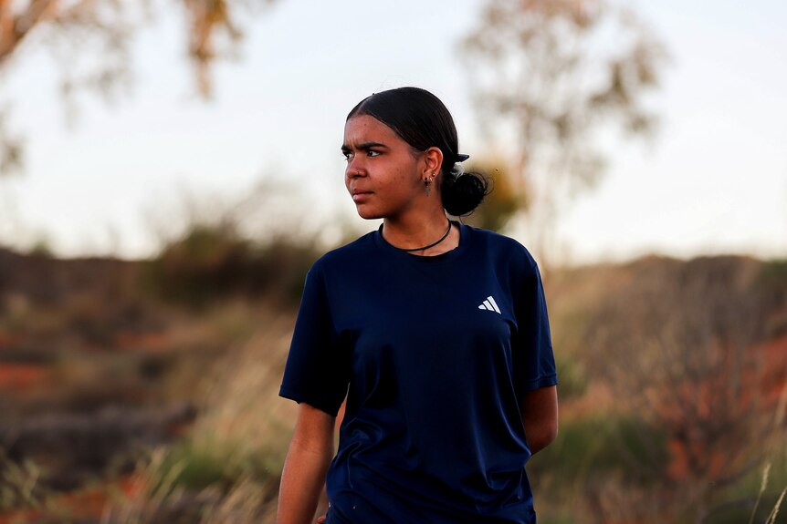 A young Aboriginal women wearing a dark blue t-shirt stands amid scrub and trees at sunset