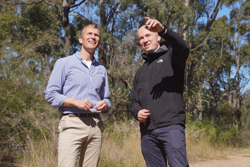 Two men, dressed casually, stand in  bushland setting with one of them pointing at trees ahead