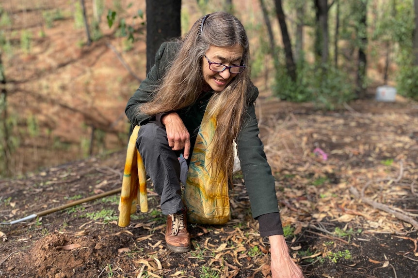 A woman kneels in bushland, pointing to the soil.