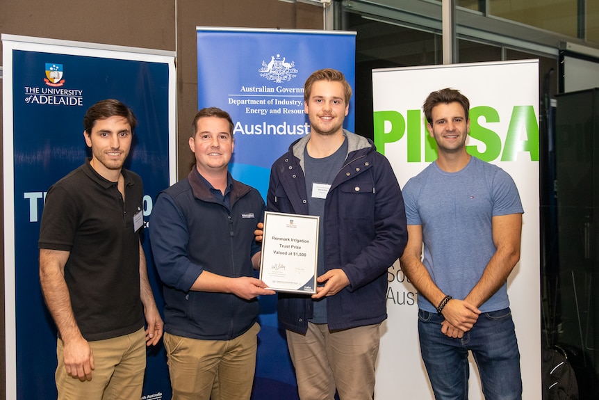 Four men stand together smiling in front of banners with an award.