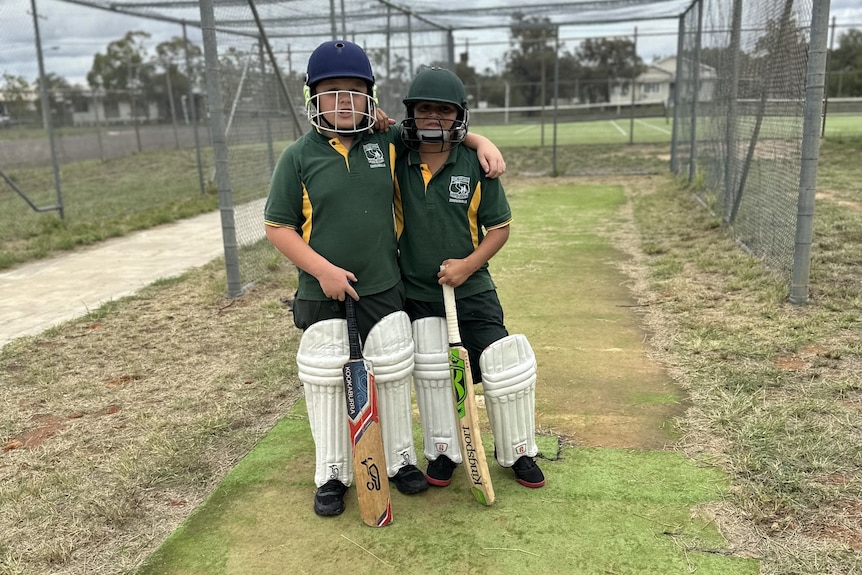 Two kids in green school informs, cricket pads, helmets and with a bat put arms around each other in cricket nets.