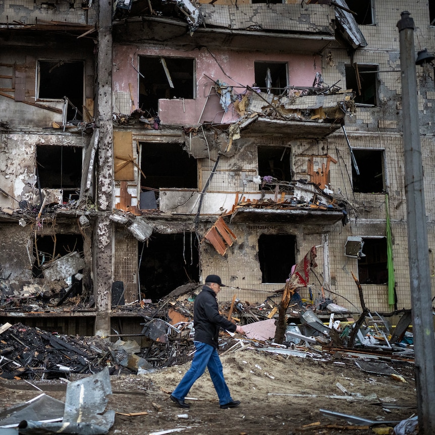 A man walks past a building damaged in a rocket attack, with wreckage strewn across the road.