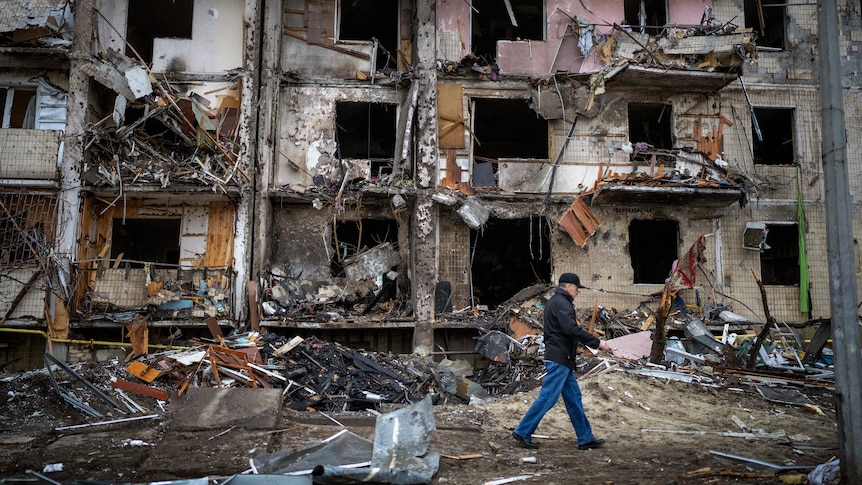 A man walks past a building damaged in a rocket attack, with wreckage strewn across the road.