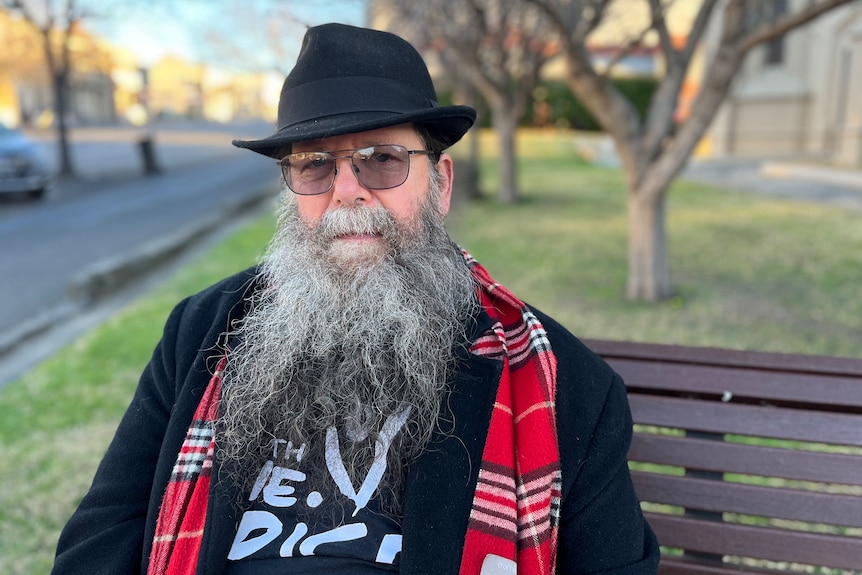 Man with long beard, black hat and red scarf sitting on a park bench