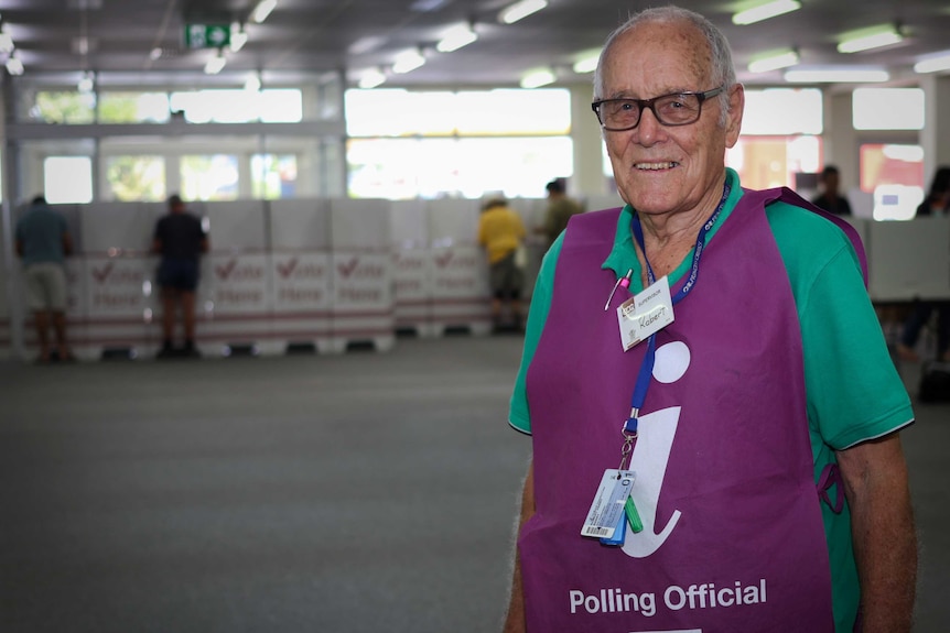 An elderly man stands inside a polling centre wearing an electoral official's vest.