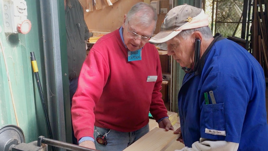 Two men stand at a woodworking station looking at what they're making and creating.
