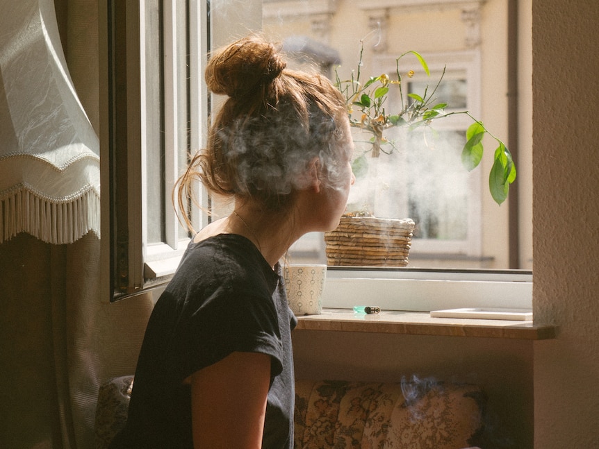 A young woman has her back to us as she looks out the window of an apartment while smoking